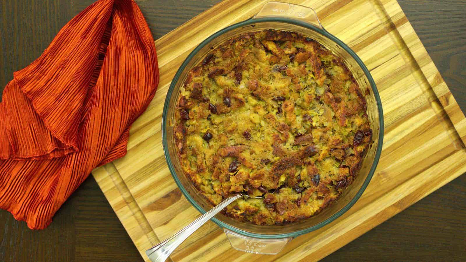 A round glass baking dish filled with golden-brown stuffing on a wooden cutting board. A silver serving spoon is placed inside. A piece of red fabric is draped to the left of the dish.