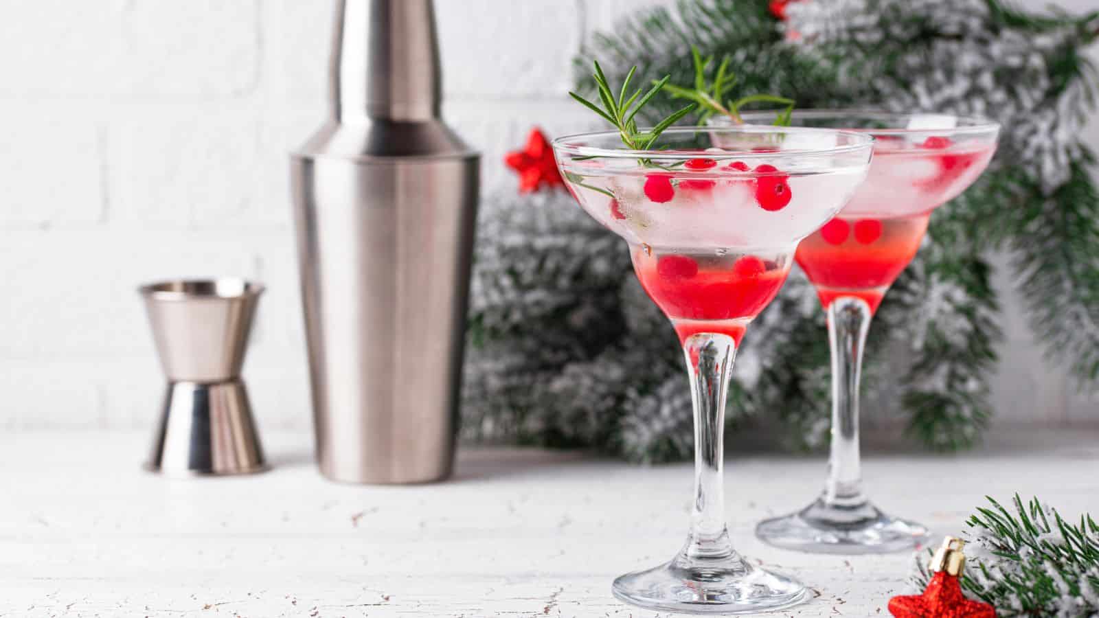 Two martini glasses filled with a clear cocktail, garnished with red berries and rosemary sprigs. A cocktail shaker and jigger sit in the background. Evergreen branches with red ornaments decorate the scene.
