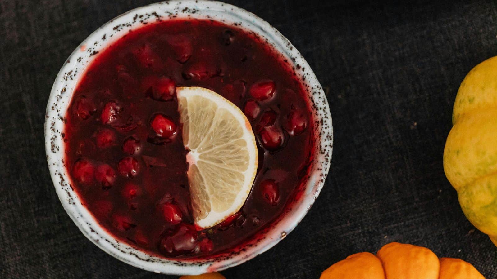 A bowl of red cranberry sauce with visible berries, topped with a slice of lemon, is placed on a dark surface. Small orange and yellow decorative gourds are partially visible nearby.