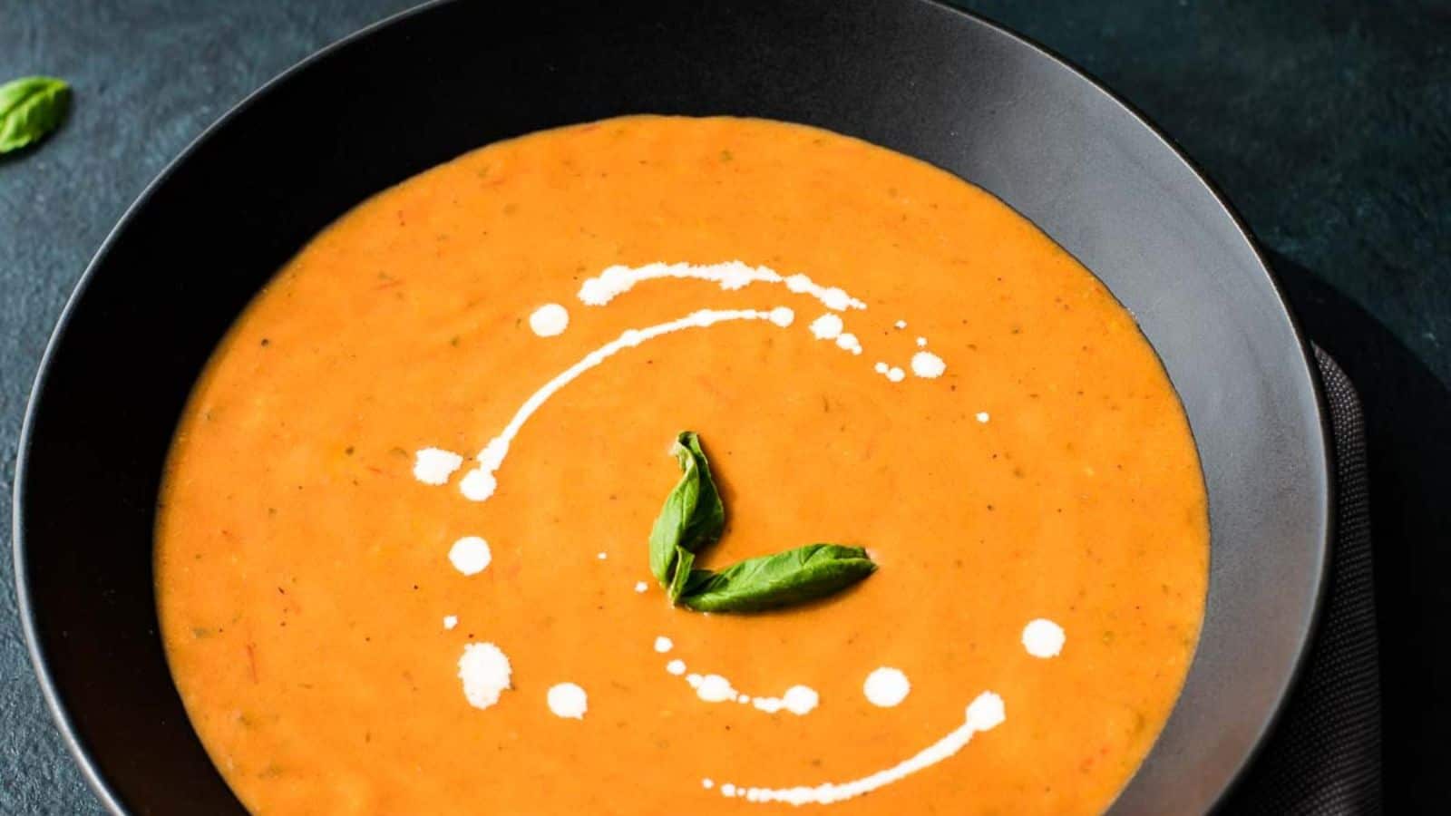 Two black bowls of tomato basil bisque on a blue background.
