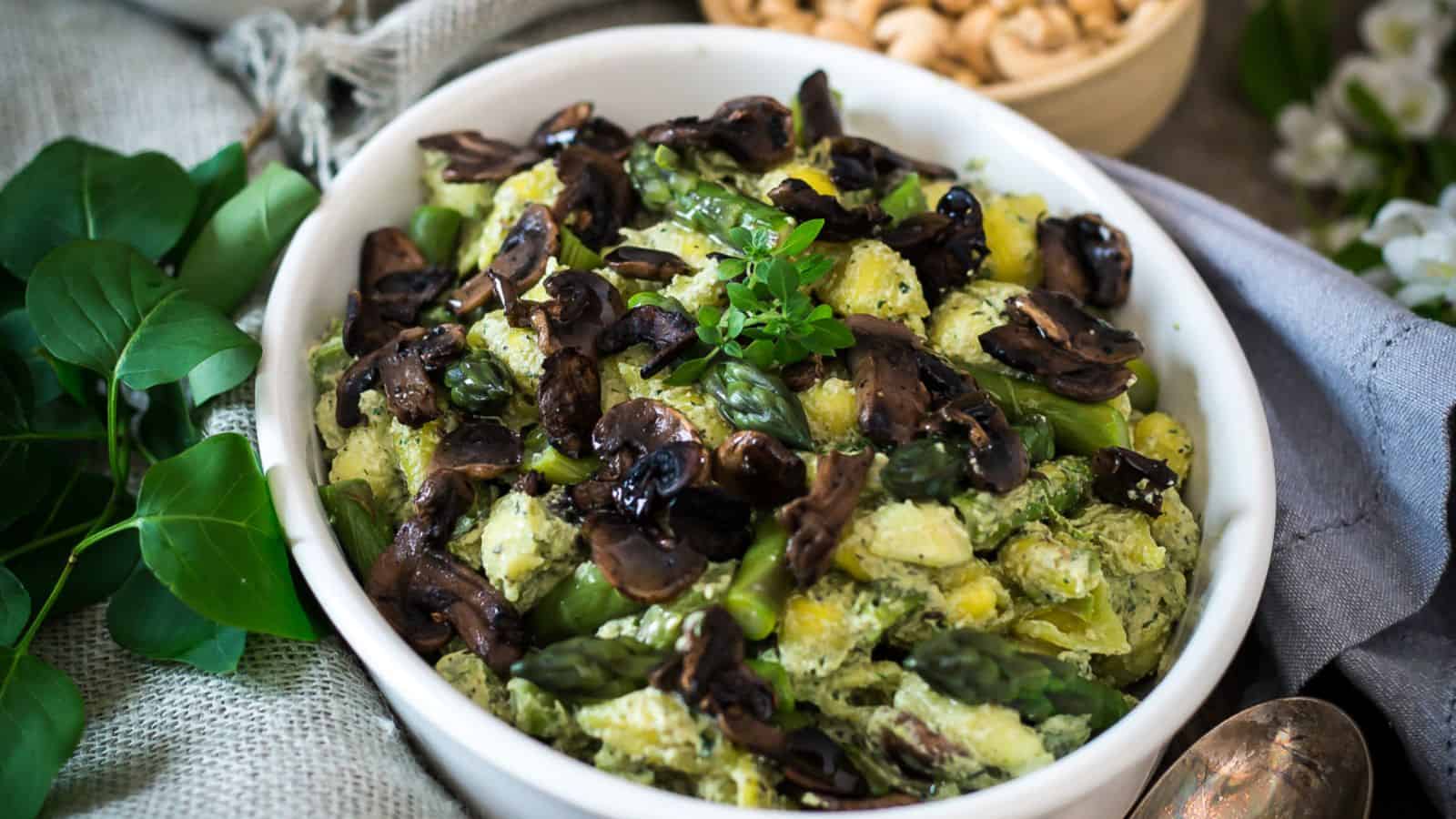 A bowl of pasta salad featuring mushrooms, asparagus, and a creamy green sauce. The dish is garnished with fresh herbs. A small bowl of nuts and some greenery are in the blurred background.