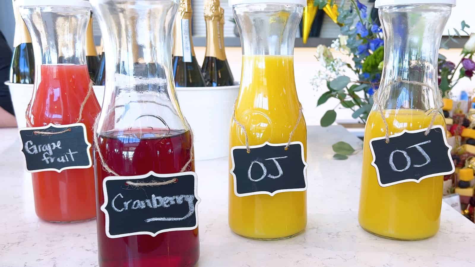 Four glass carafes are filled with different juices on a countertop. The drinks include grapefruit juice, cranberry juice, and two carafes of orange juice, each labeled with a small chalkboard sign. Wine bottles and flowers are seen in the background.