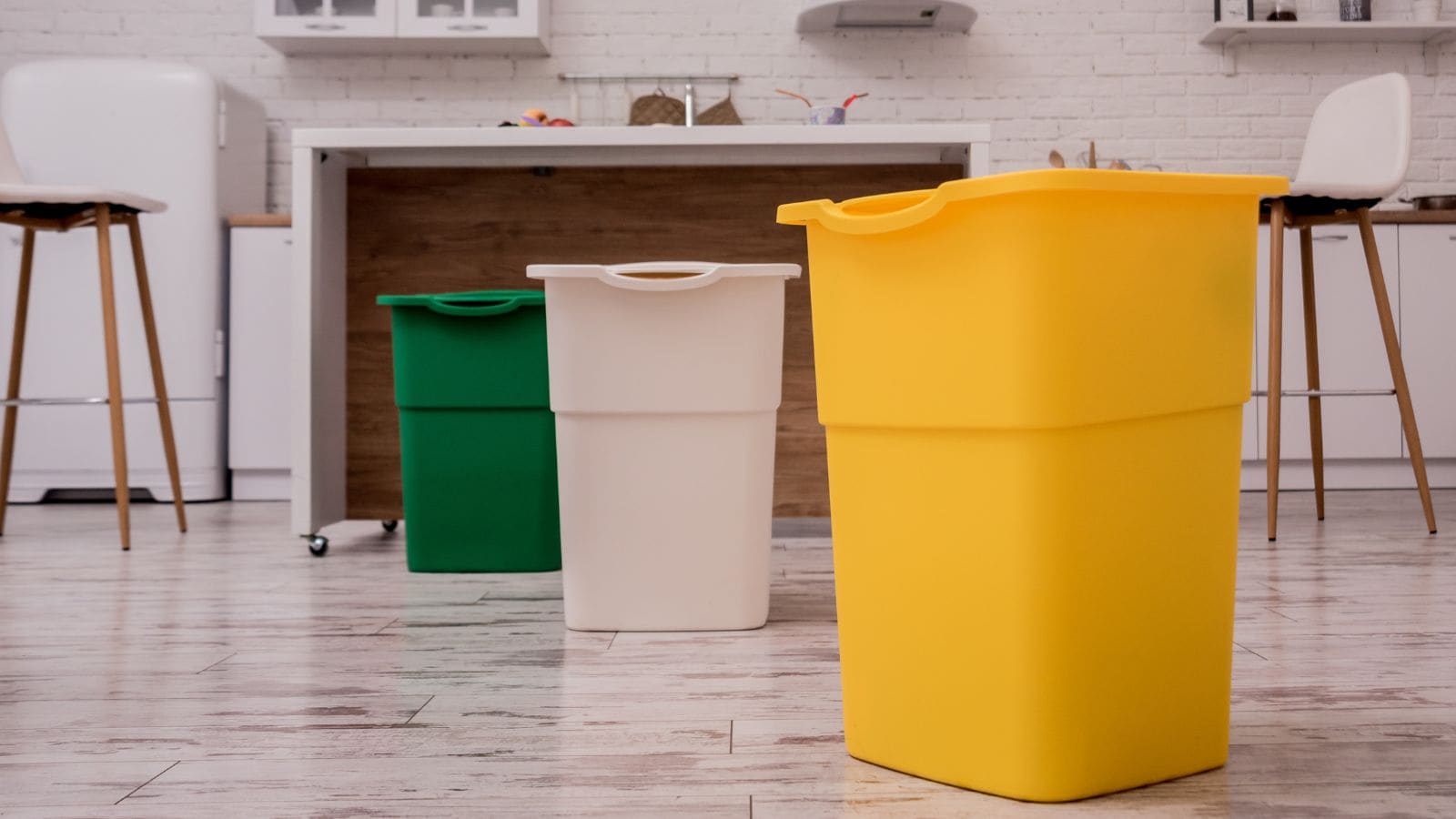 A kitchen with three tall recycling bins in yellow, white, and green on a tiled floor. The background features a small table with two high chairs, a cupboard, and a white brick wall.