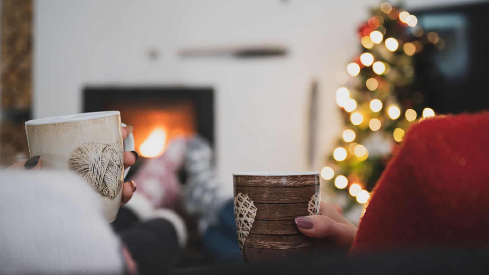 Two people relaxing by a warm fireplace, holding mugs of hot beverages with a Christmas tree softly glowing in the background.