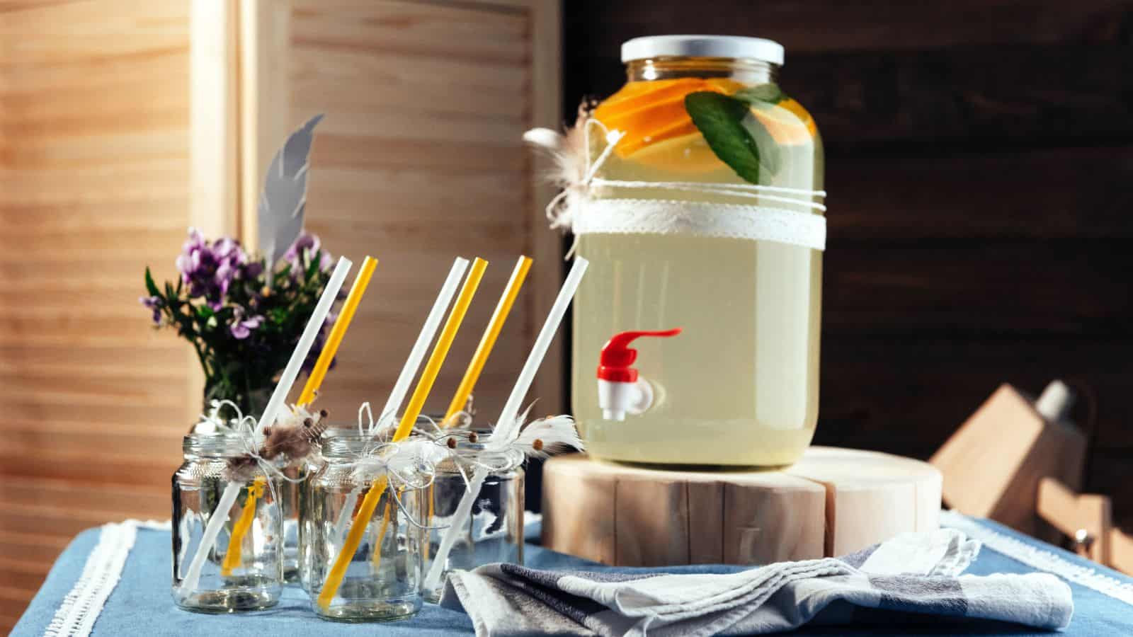 A rustic self-serve drink station featuring a glass dispenser of citrus-infused water and decorative jars with straws on a blue tablecloth.