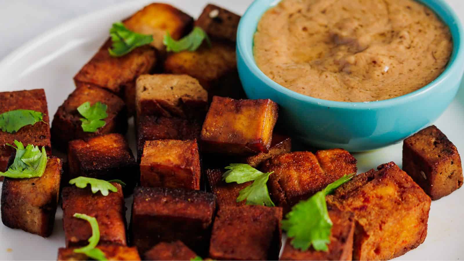Crispy tofu cubes garnished with cilantro are arranged on a plate next to a small blue bowl filled with a creamy brown dipping sauce.