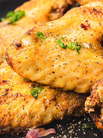 Close-up of seasoned and grilled chicken wings garnished with chopped herbs on a black plate. Lemon wedges are visible in the background, adding a touch of color.
