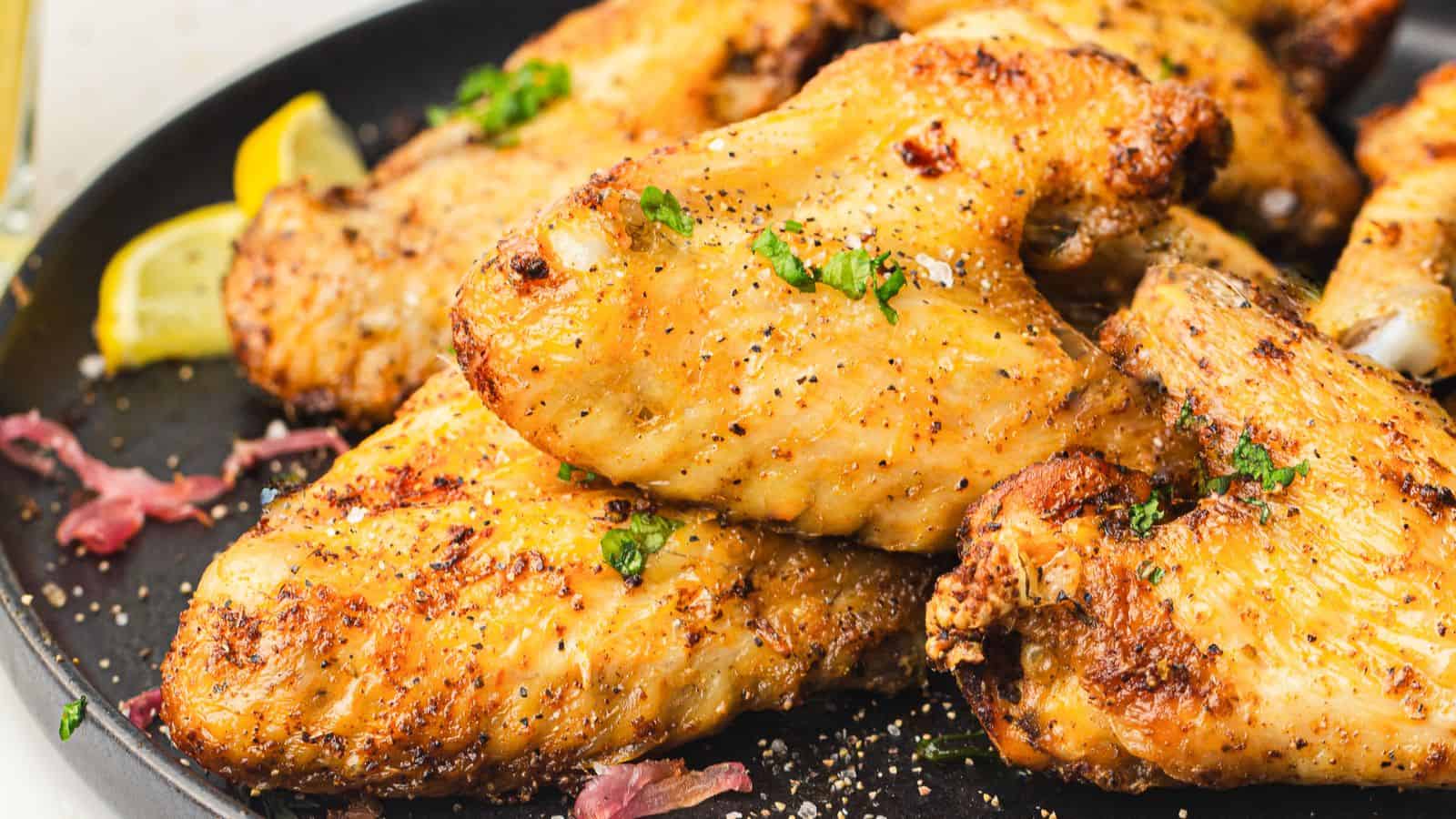 Close-up of a plate of seasoned and grilled chicken wings garnished with herbs. Lemon wedges are visible in the background, adding a hint of color. The wings appear crispy and well-cooked, resting on a dark plate.