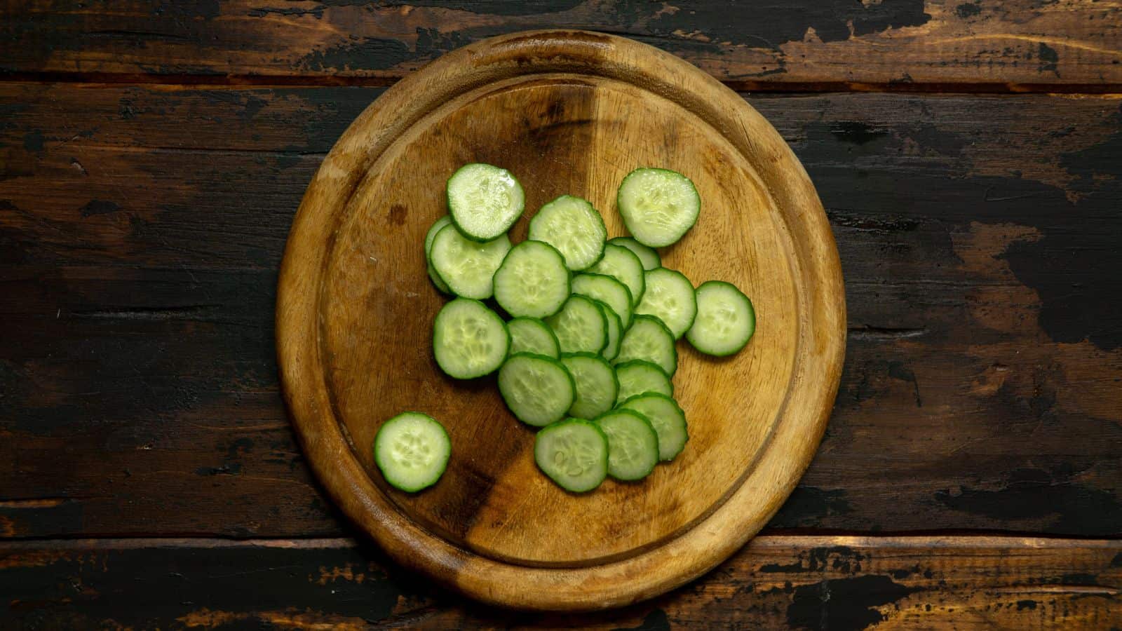 A wooden round cutting board with several slices of cucumber on it, placed on a dark wooden table.