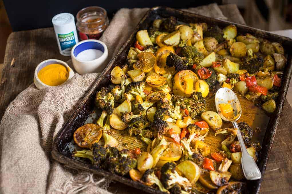 A roasted vegetable sheet pan on a rustic table includes potatoes, broccoli, and peppers. A spoon rests on the pan. Nearby are small bowls of turmeric, salt, and a glass jar with spices. A burlap cloth is partially visible underneath.