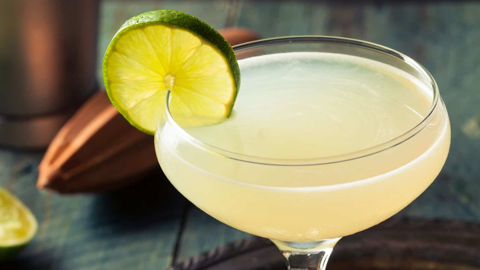A close-up of a cocktail in a coupe glass garnished with a lime slice. The drink is light yellow and appears smooth. A wooden citrus juicer and a halved lime are visible in the blurred background.