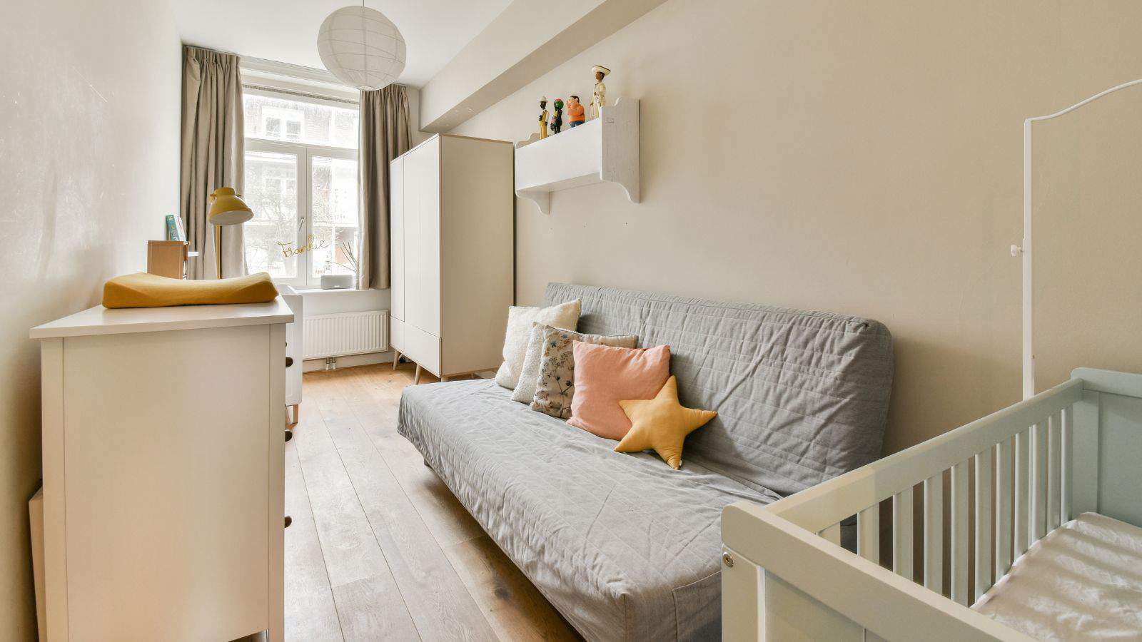 A cozy nursery room features a gray futon with colorful cushions, a white dresser with a changing pad, and a light-colored crib. A shelf with toys is mounted above the futon. Natural light streams in through a window with sheer curtains.