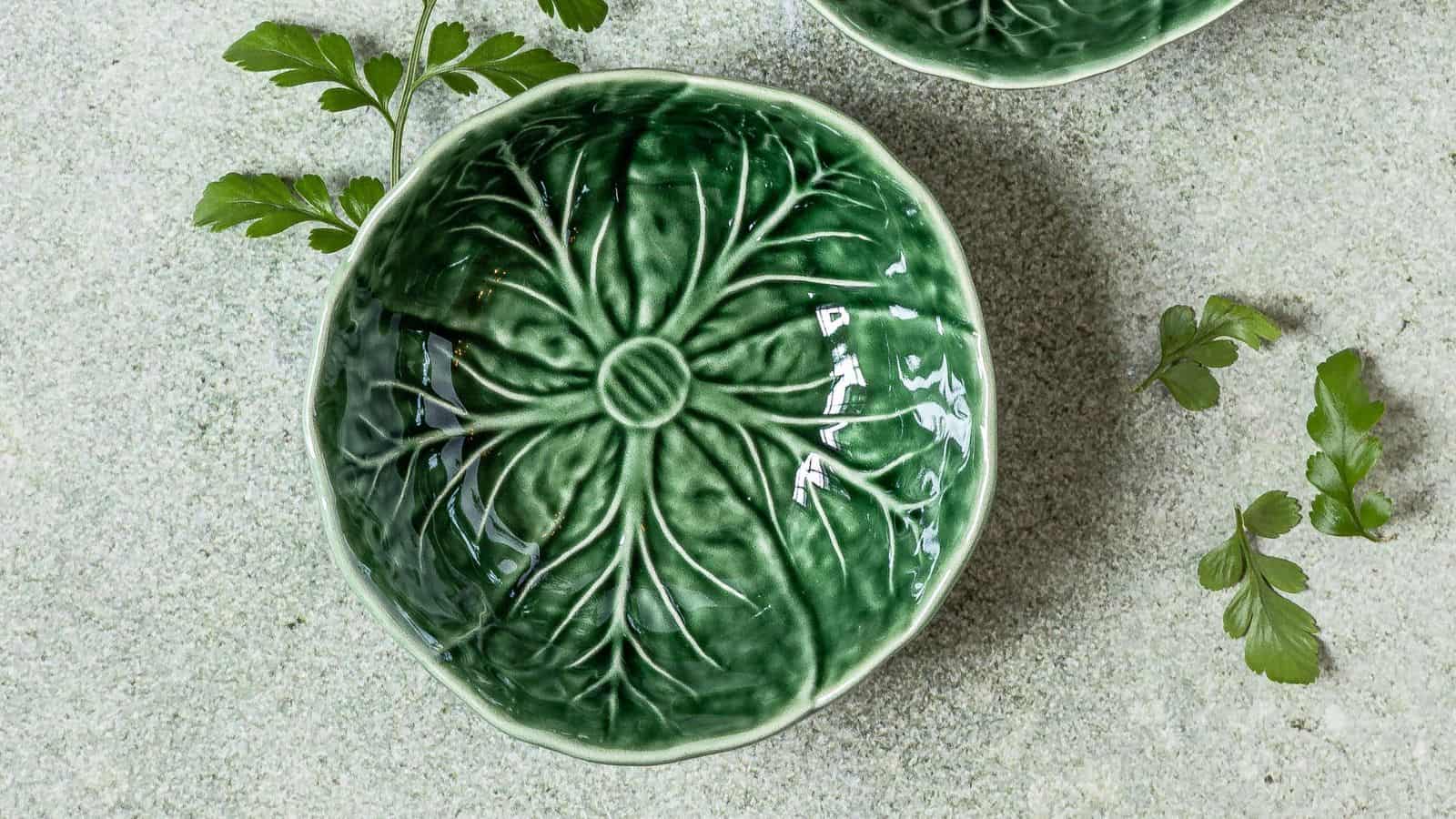 A textured green ceramic bowl with a leaf pattern on the inside is placed on a gray surface. The bowl is accompanied by sprigs of green leaves scattered nearby.
