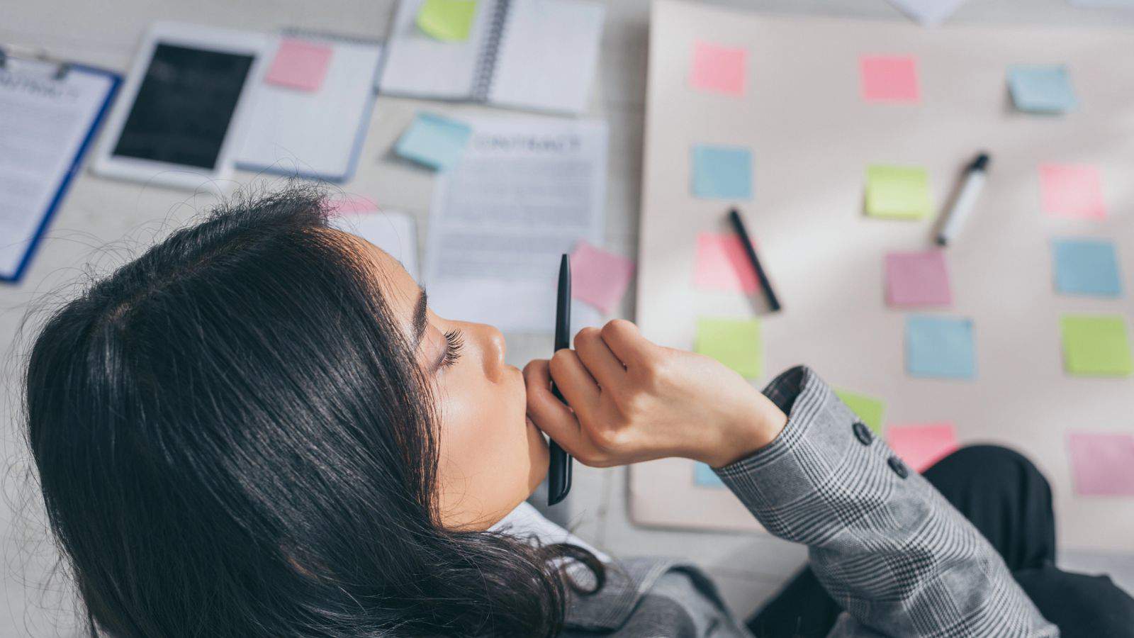 A woman sits on the floor, holding a pen near her chin, surrounded by scattered papers, notebooks, and a large pad covered with colorful sticky notes. She appears deep in thought while organizing her notes.