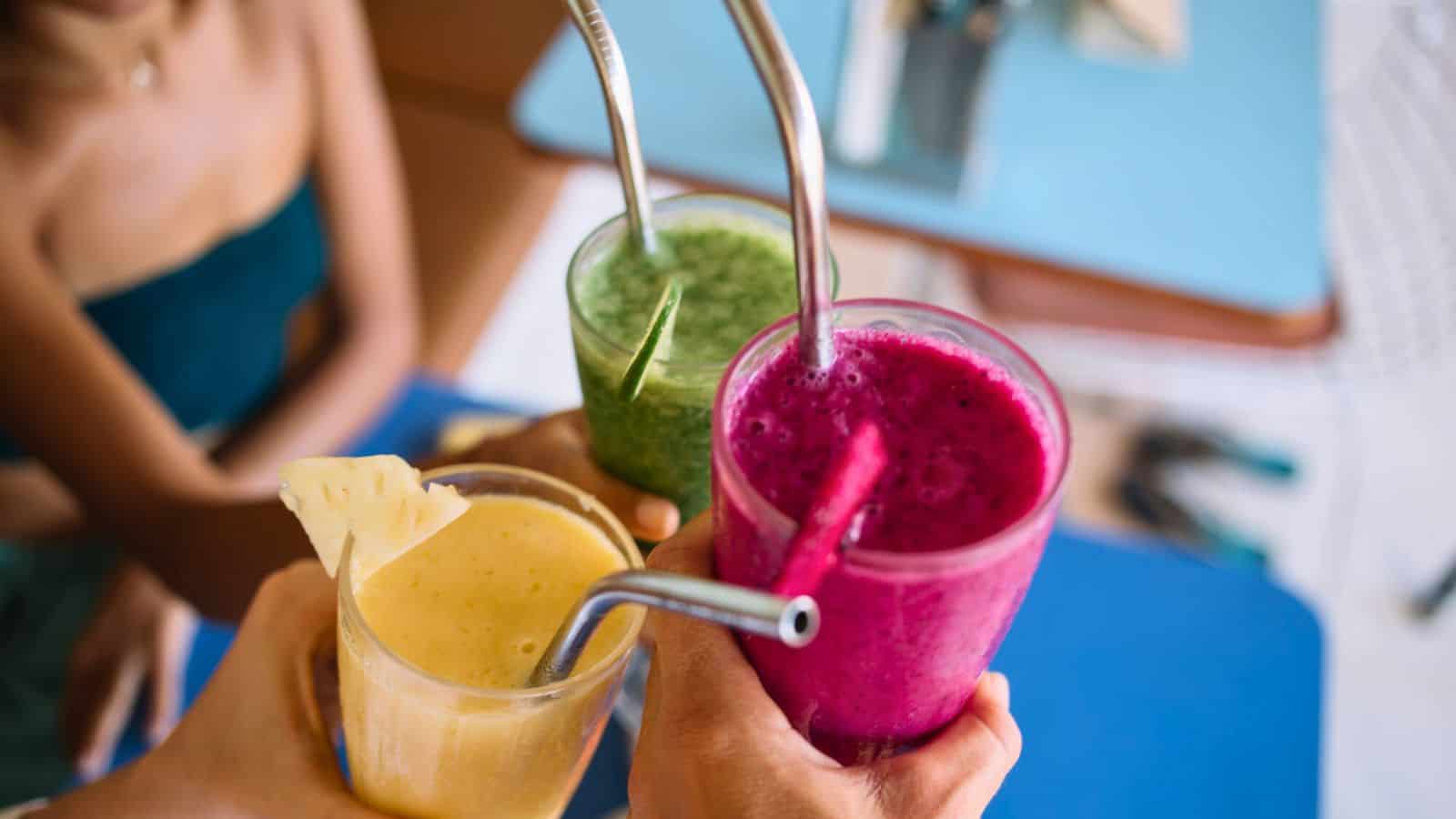 Three hands holding colorful smoothies with metal straws are raised in a toast. The smoothies are green, purple, and yellow, each garnished with a fruit slice. A blurred person is in the background, and a blue surface is partially visible.