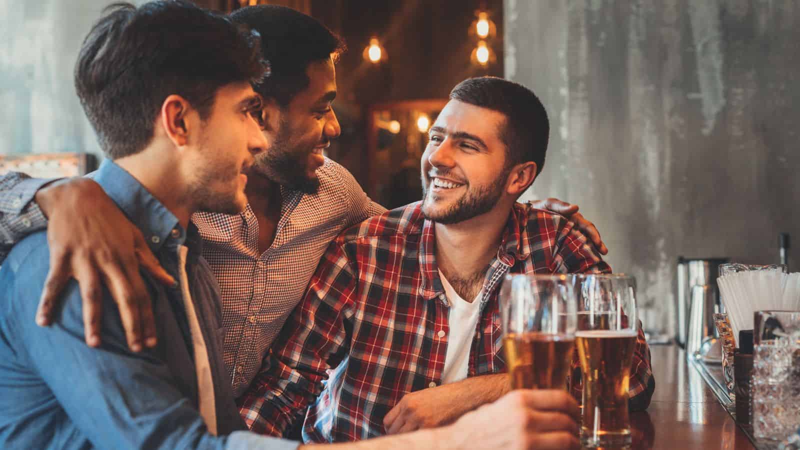 Three men sit at a bar, each with a glass of beer. They are smiling and engaged in conversation. The bar has a warm ambiance with soft lighting and a row of drinks on the counter.