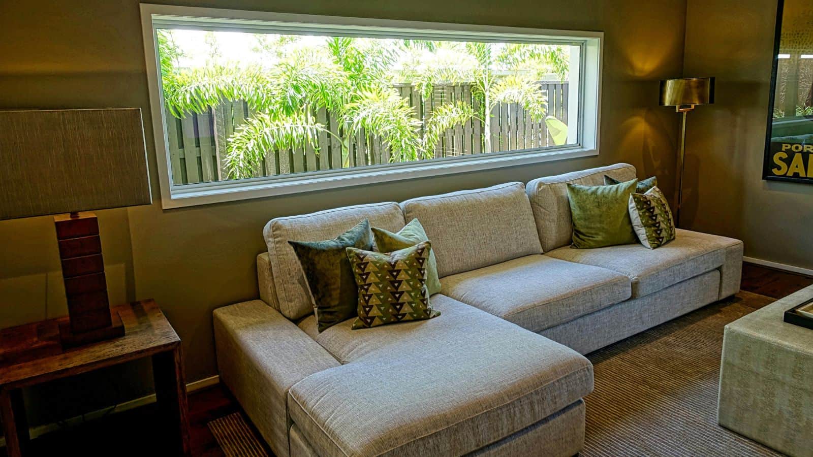 A modern living room with a large beige sectional sofa topped with green and patterned pillows. A wooden side table holds a lamp with a square shade. A long horizontal window reveals greenery outside. The room has neutral-colored walls and carpet.