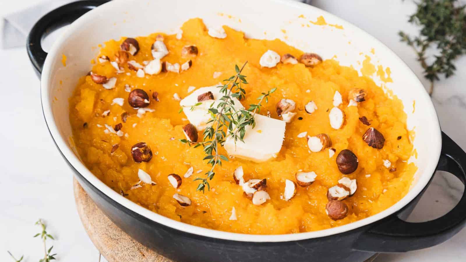 A dish filled with mashed sweet potatoes topped with chopped nuts, a pat of butter, and a sprig of thyme. The dish is placed on a wooden board, with a few thyme sprigs scattered around.