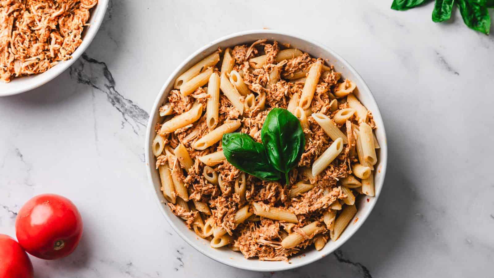 A bowl of penne pasta topped with shredded meat and garnished with fresh basil leaves. A fork is placed in the bowl. Red tomatoes and additional fresh basil leaves are on the marble surface nearby.