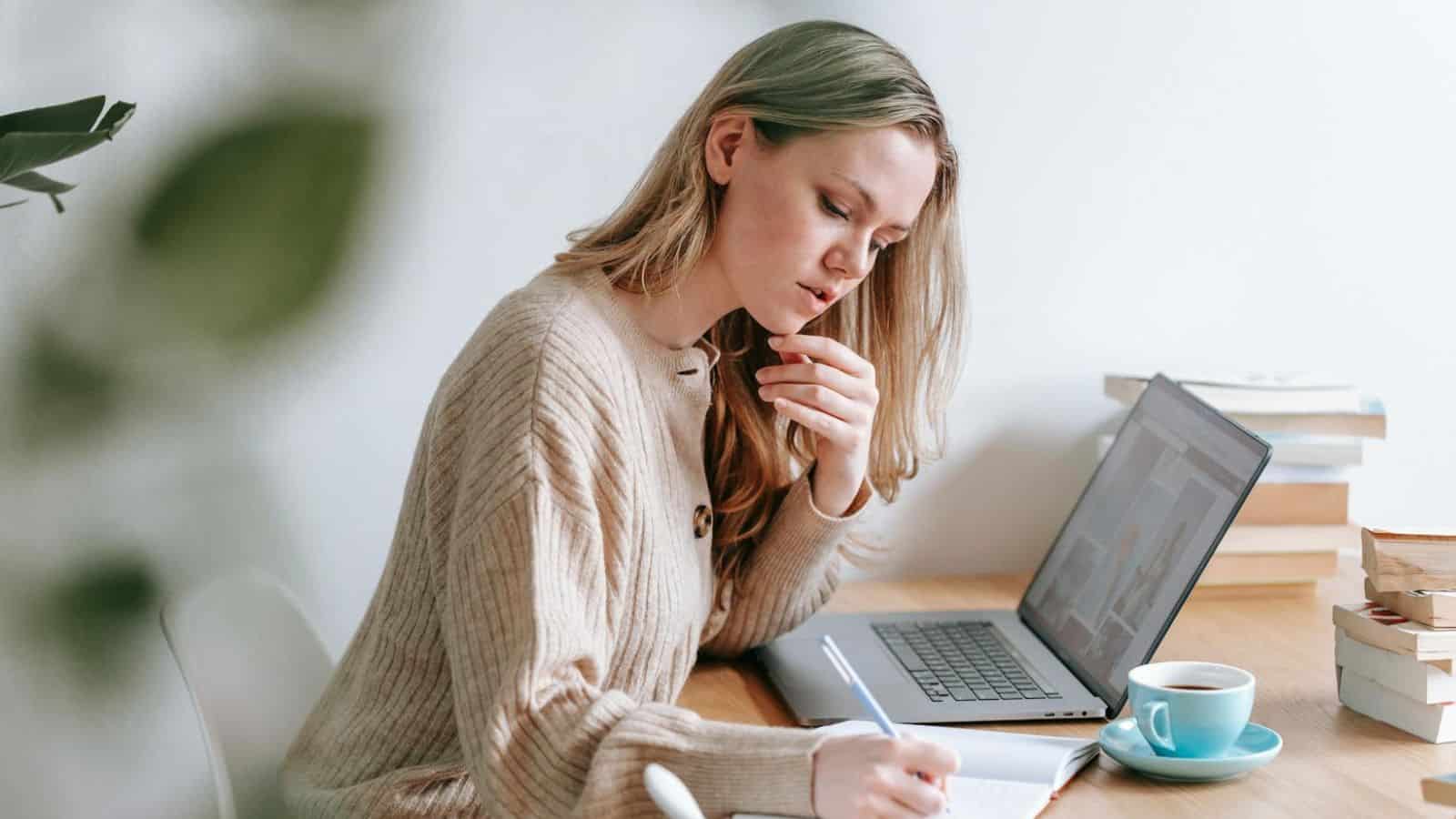 A woman with long hair sits at a wooden table, writing in a notebook with a pen. She is wearing a beige sweater and focusing intently. A laptop is open next to her, and a blue cup filled with a beverage is on the table.