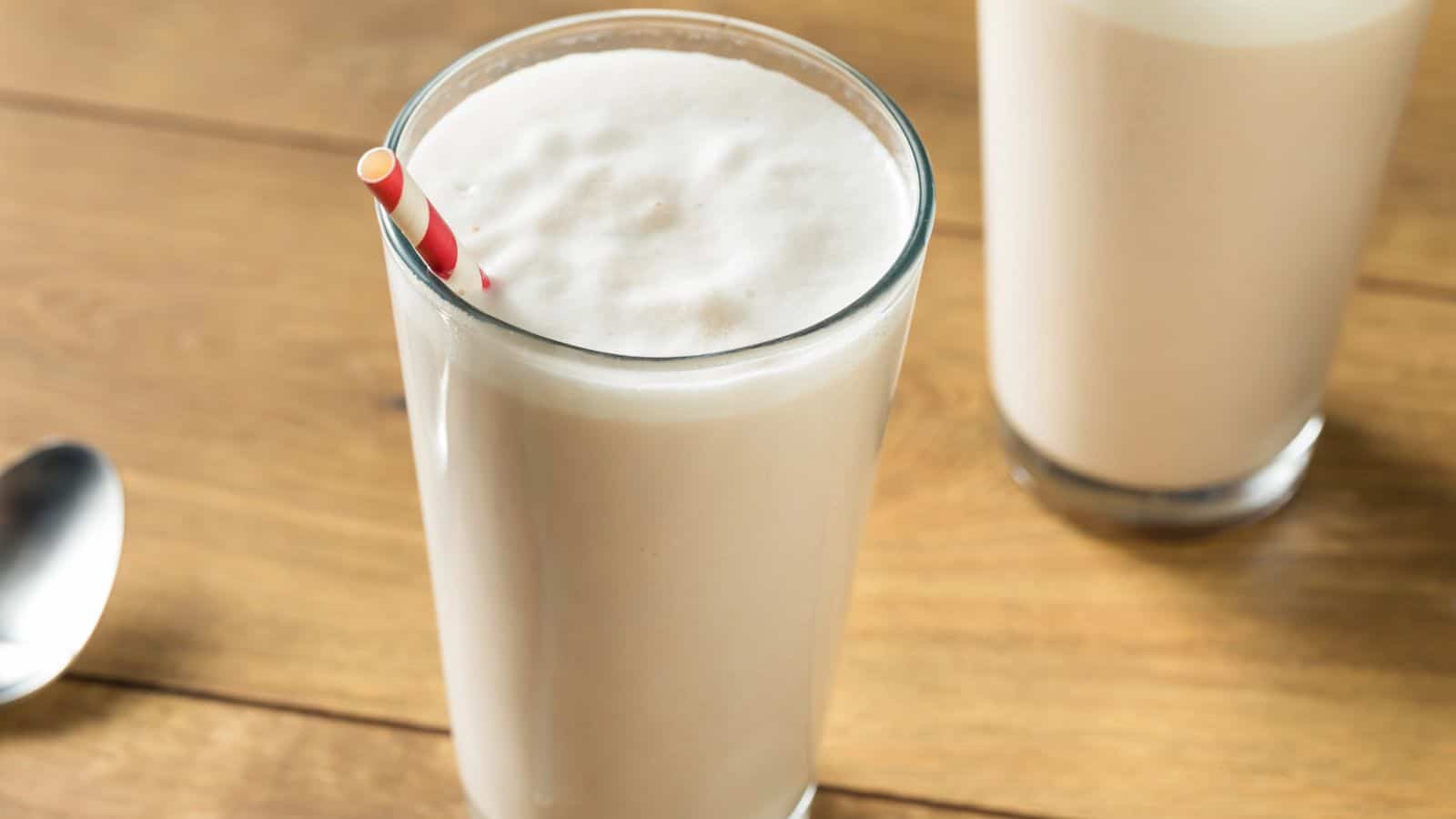 A glass of vanilla milkshake with a red and white striped straw sits on a wooden surface. Another glass is partially visible in the background. A silver spoon rests to the left.