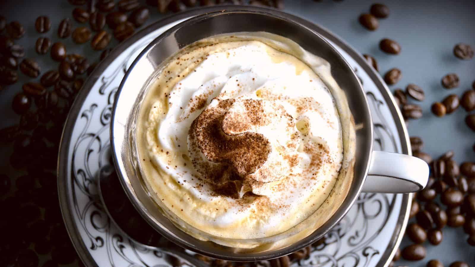 A cup of coffee topped with whipped cream and a sprinkle of cocoa powder, placed on a decorative saucer. Coffee beans are scattered around the cup on the surface.
