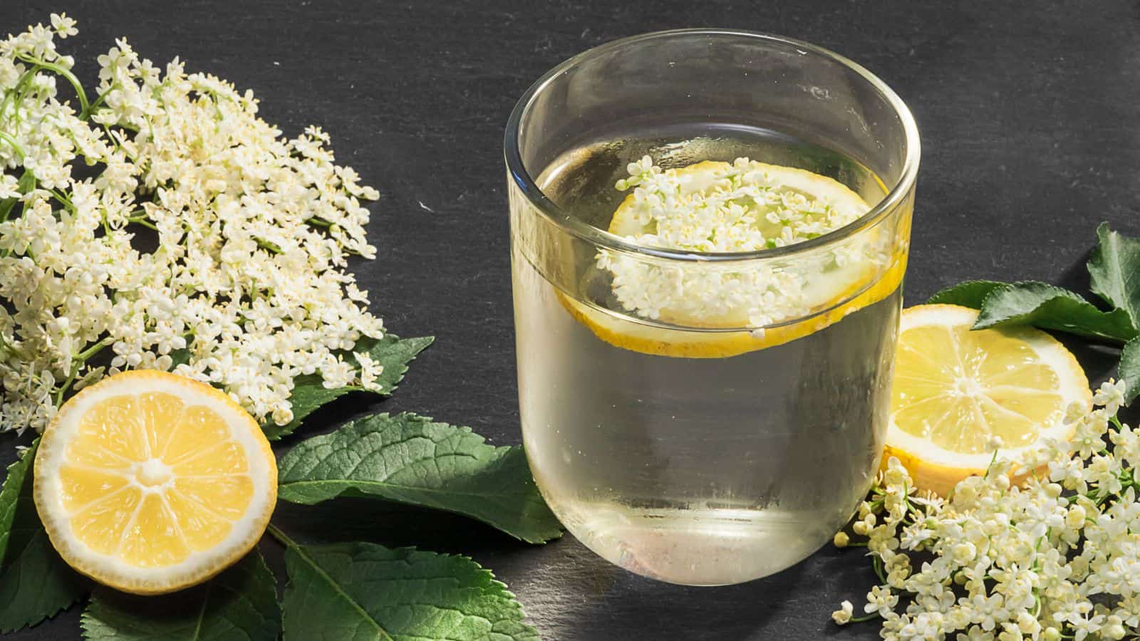 A glass of elderflower lemonade is garnished with a lemon slice and elderflowers, surrounded by fresh elderflowers, lemon slices, and green leaves on a dark surface.