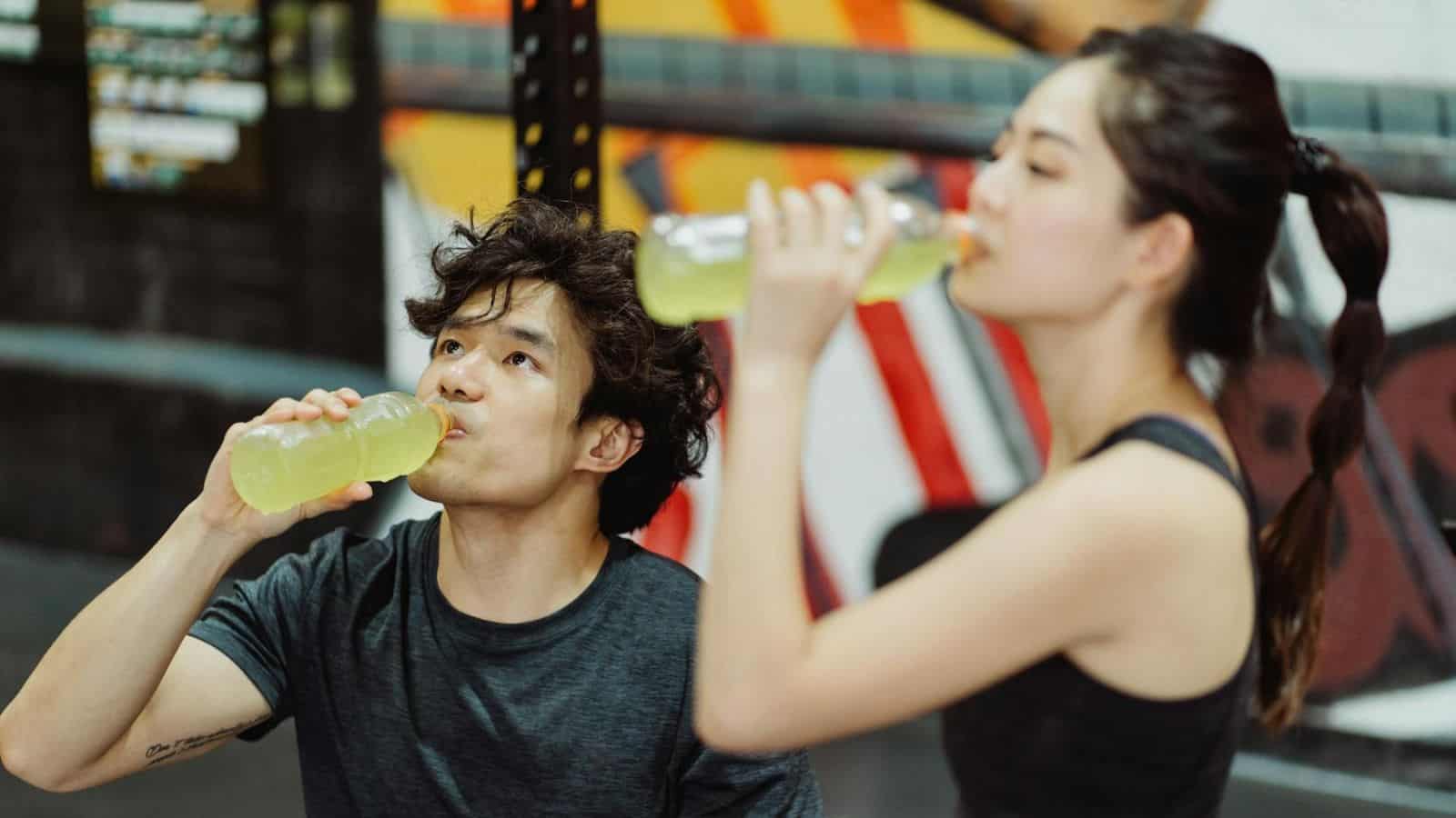 A man and a woman are sitting in a gym and drinking from bottles with a light green beverage. They appear to be taking a break from exercising. The background features gym equipment and a partially visible colorful wall mural.