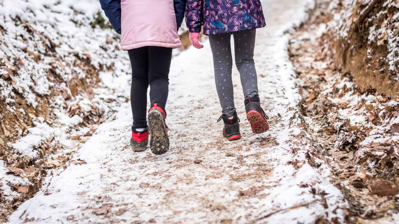 Two people, wearing winter jackets and leggings, walk side by side on a snowy path. The ground is covered with patches of snow and leaves. The focus is on their legs and footwear.