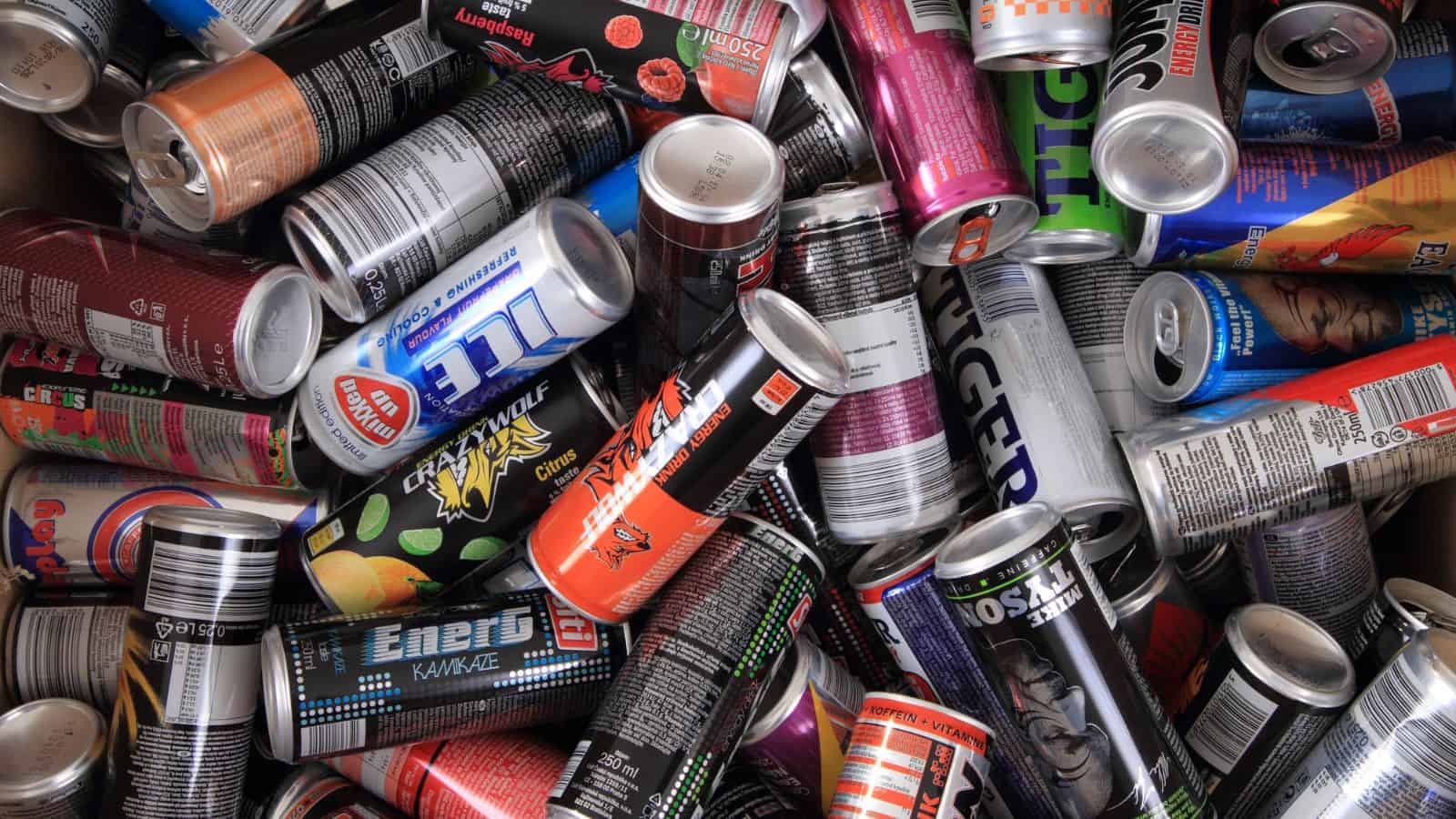 A collection of various energy drink cans in different colors and designs, scattered in a pile. The brands and labels are partially visible, featuring a mix of bright and dark shades, contributing to a visually busy scene.