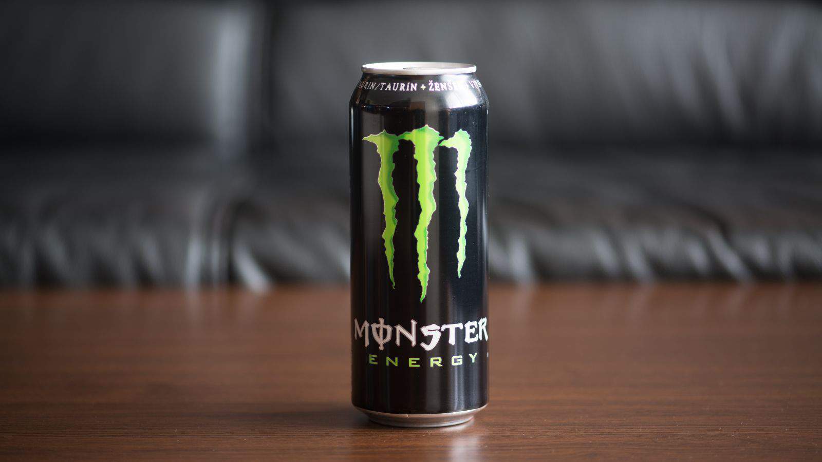 A can of Monster Energy drink sits on a wooden surface with a black leather couch in the background. The can is predominantly black with the green Monster logo prominently displayed in the center.
