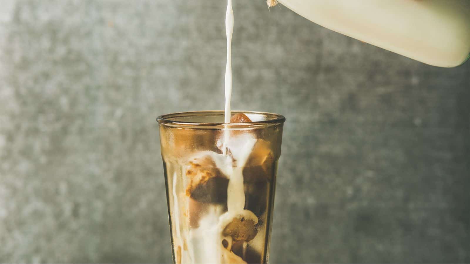 Milk is being poured from a container into a glass filled with ice cubes, creating a creamy swirl. The background is a muted gray.