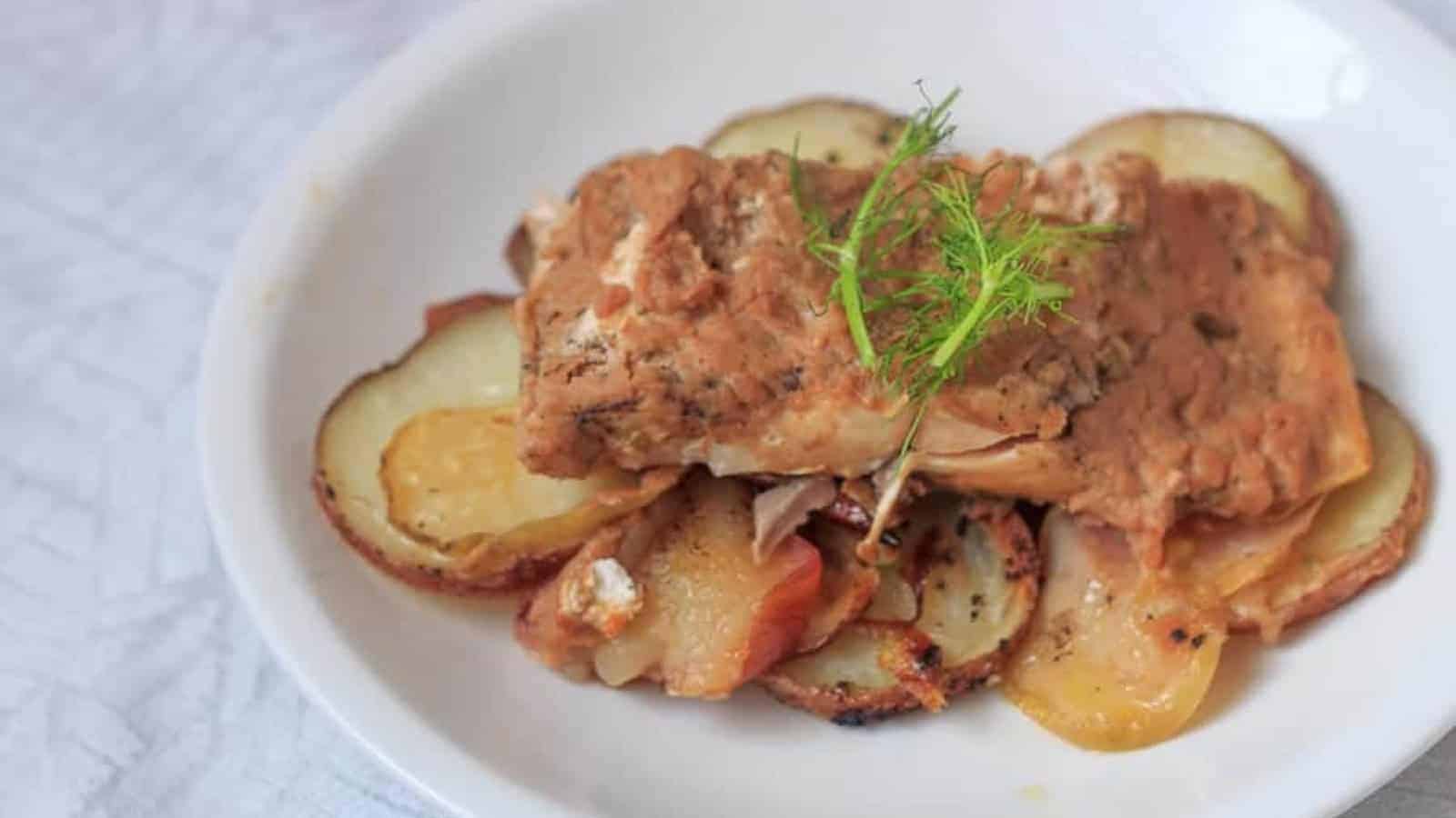 A plate of cooked sliced potatoes topped with a piece of seasoned salmon and garnished with a sprig of dill. The dish is presented on a white plate with a subtle textured background.