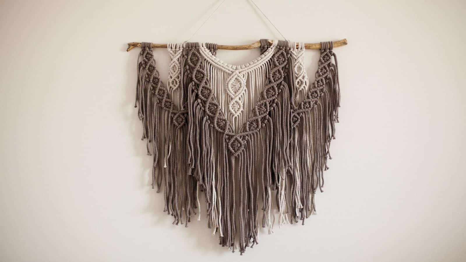 A handcrafted macram&eacute; wall hanging features intricate knots in a symmetrical pattern. It is made from beige and brown cords, with long, fringed tassels hanging from a wooden dowel. The piece is mounted against a plain white wall.