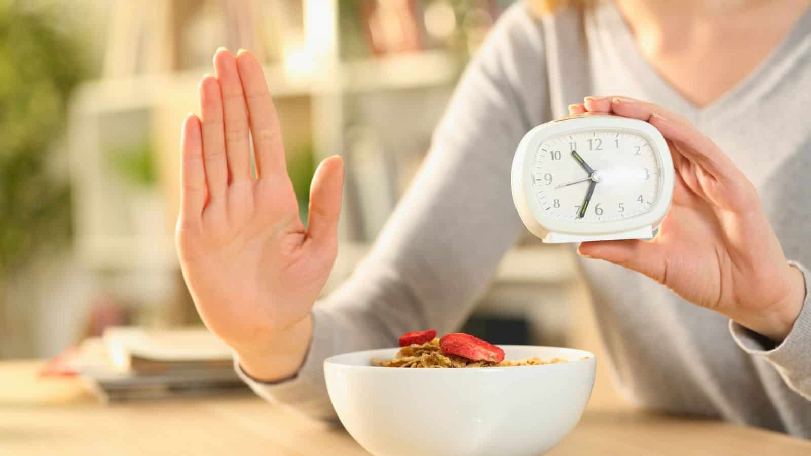 A person holds up a small clock with one hand, displaying 7:00, while extending their other hand in a gesture of refusal in front of a bowl of cereal topped with sliced strawberries on a table.