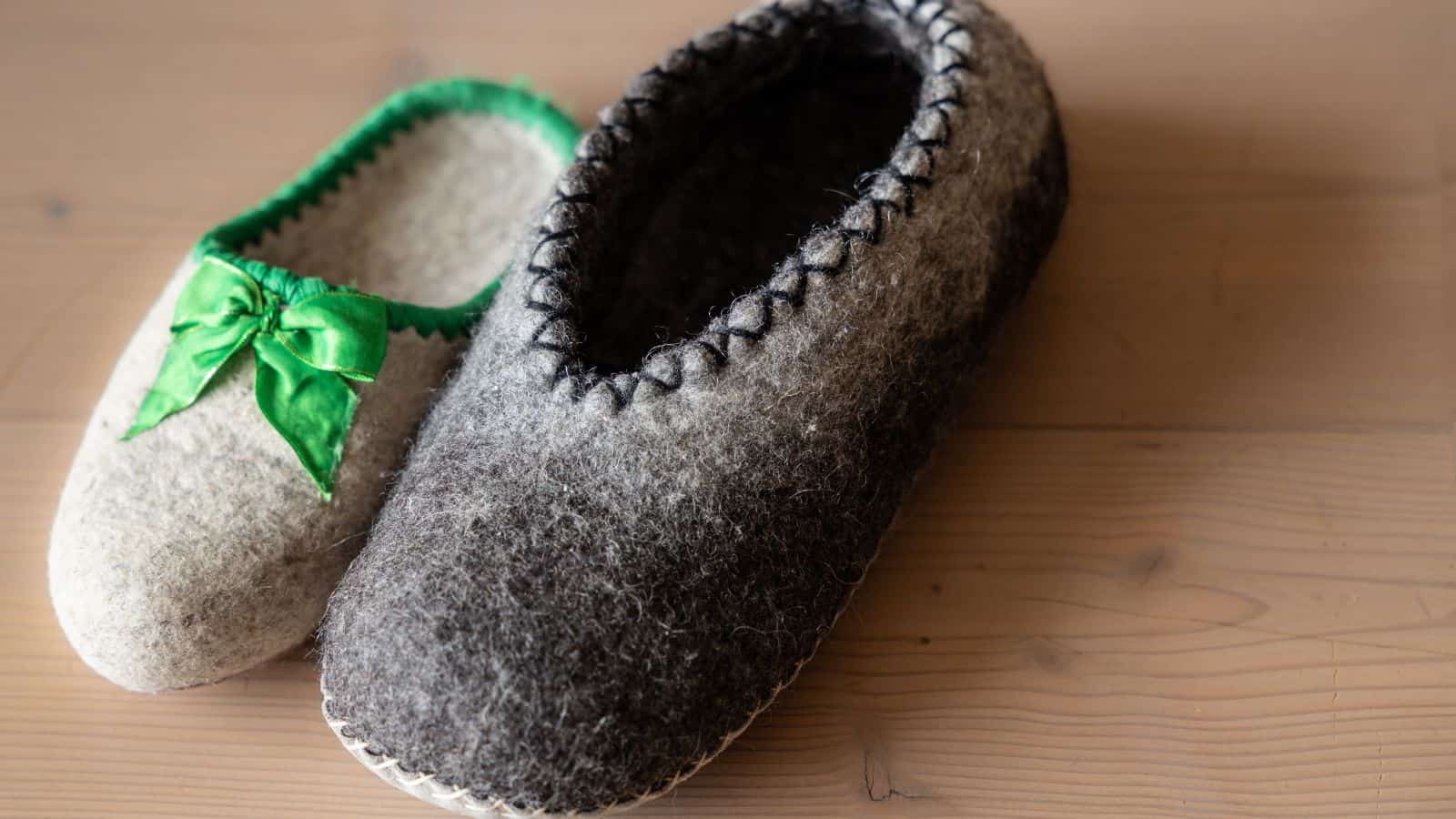 Two woolen slippers rest on a wooden surface, perfect for cozy winter DIY projects. The left slipper is light gray with a green bow, while the right is dark gray with black stitching around the opening. Both slippers showcase a charming handmade touch.