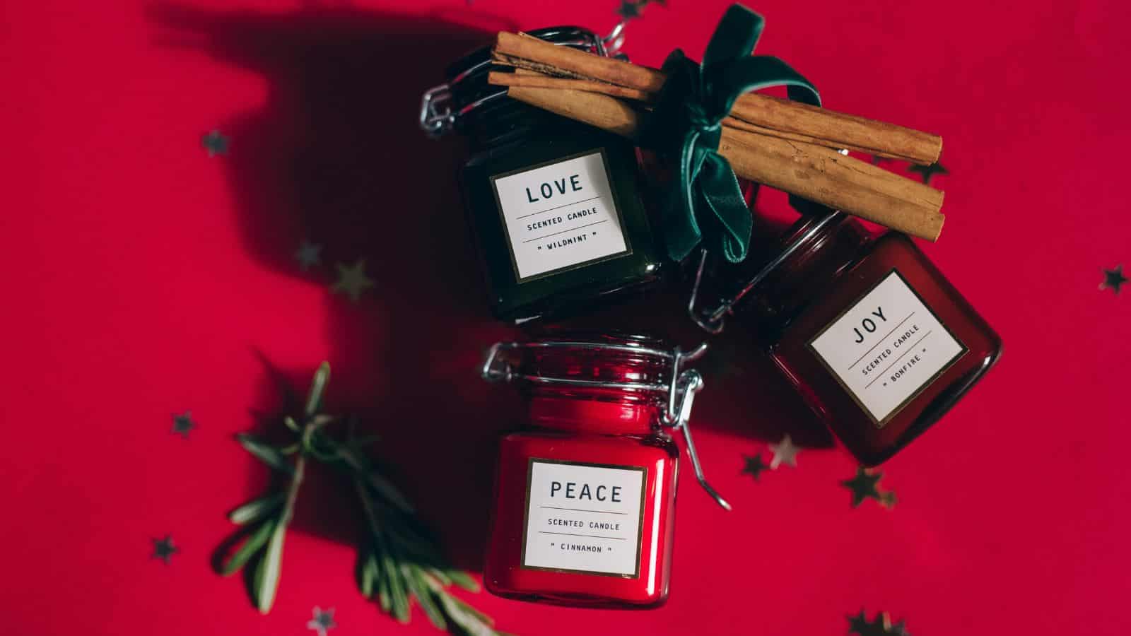 Three scented candles labeled "Love," "Peace," and "Joy" are placed on a red background. The candles are red and dark green, and tied together with a ribbon. Cinnamon sticks and rosemary are arranged nearby, with star-shaped confetti scattered around.