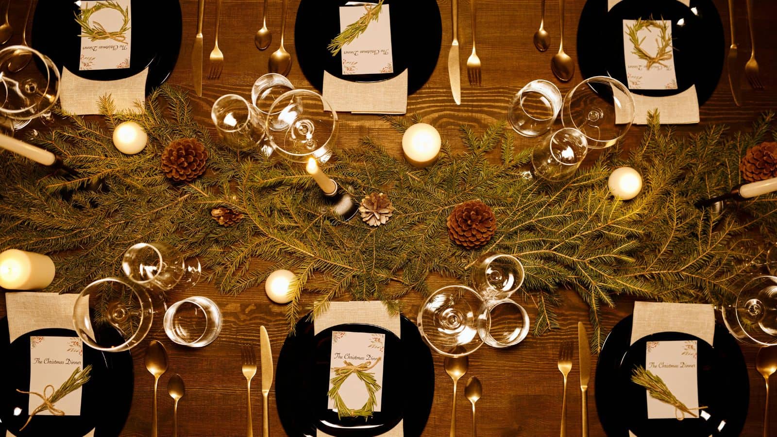A wooden dining table is set with black plates, silver cutlery, and glassware. Menus rest on white napkins. The centerpiece features greenery, pinecones, and lit candles, creating a festive atmosphere.