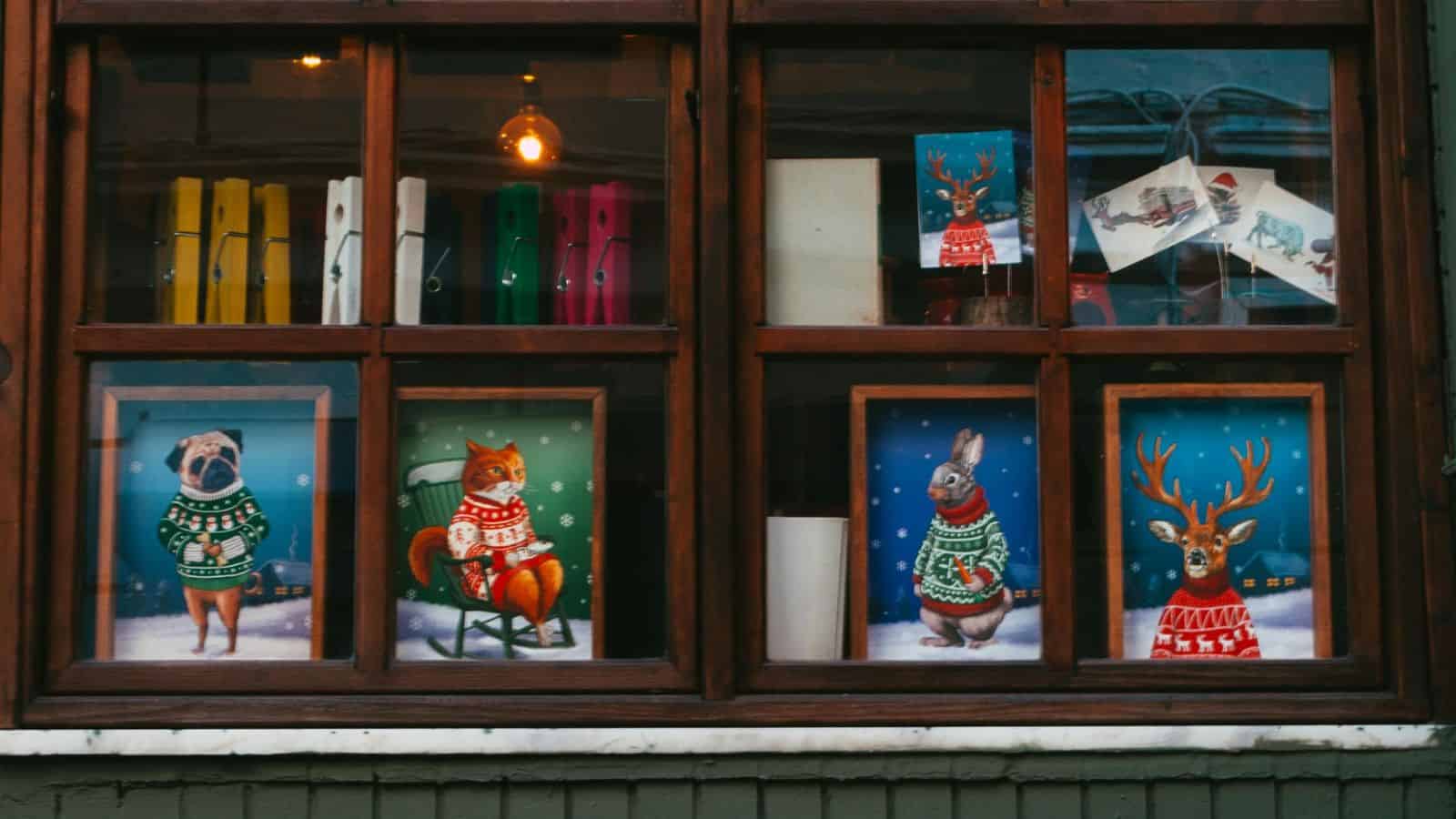A window display with framed pictures of animals in Christmas sweaters, including a pug, fox, rabbit, and deer. Colorful books are on the left, and holiday-themed cards are on the upper right. A warm light bulb illuminates the scene.