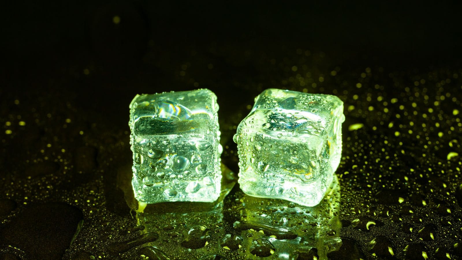 Close-up of two clear ice cubes with trapped bubbles, illuminated with green light, creating a dramatic effect against a dark background.