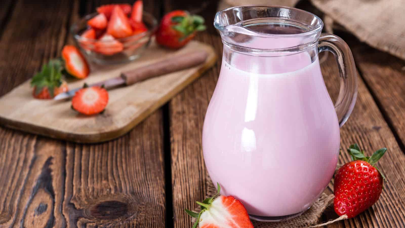 A glass pitcher filled with pink strawberry milk is placed on a wooden table. Fresh strawberries and a wooden cutting board with sliced strawberries are in the background. A knife rests on the board beside a glass bowl.
