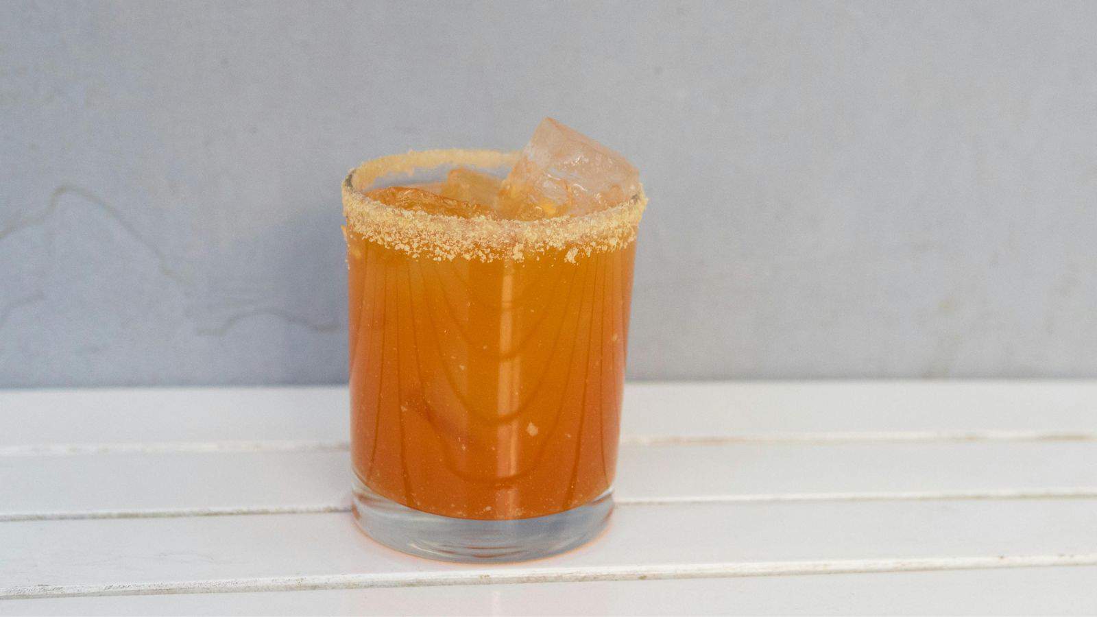 Amber-colored drink in a glass with a rim coated in spiced salt, with large ice cubes floating in the beverage, placed on a white surface.