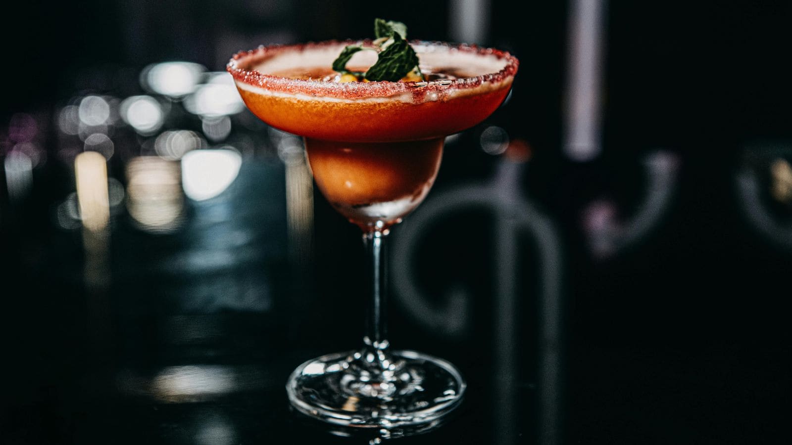 Dark cocktail in a margarita glass with a red sugar-coated rim, garnished with fresh mint leaves, standing against a dimly lit background.