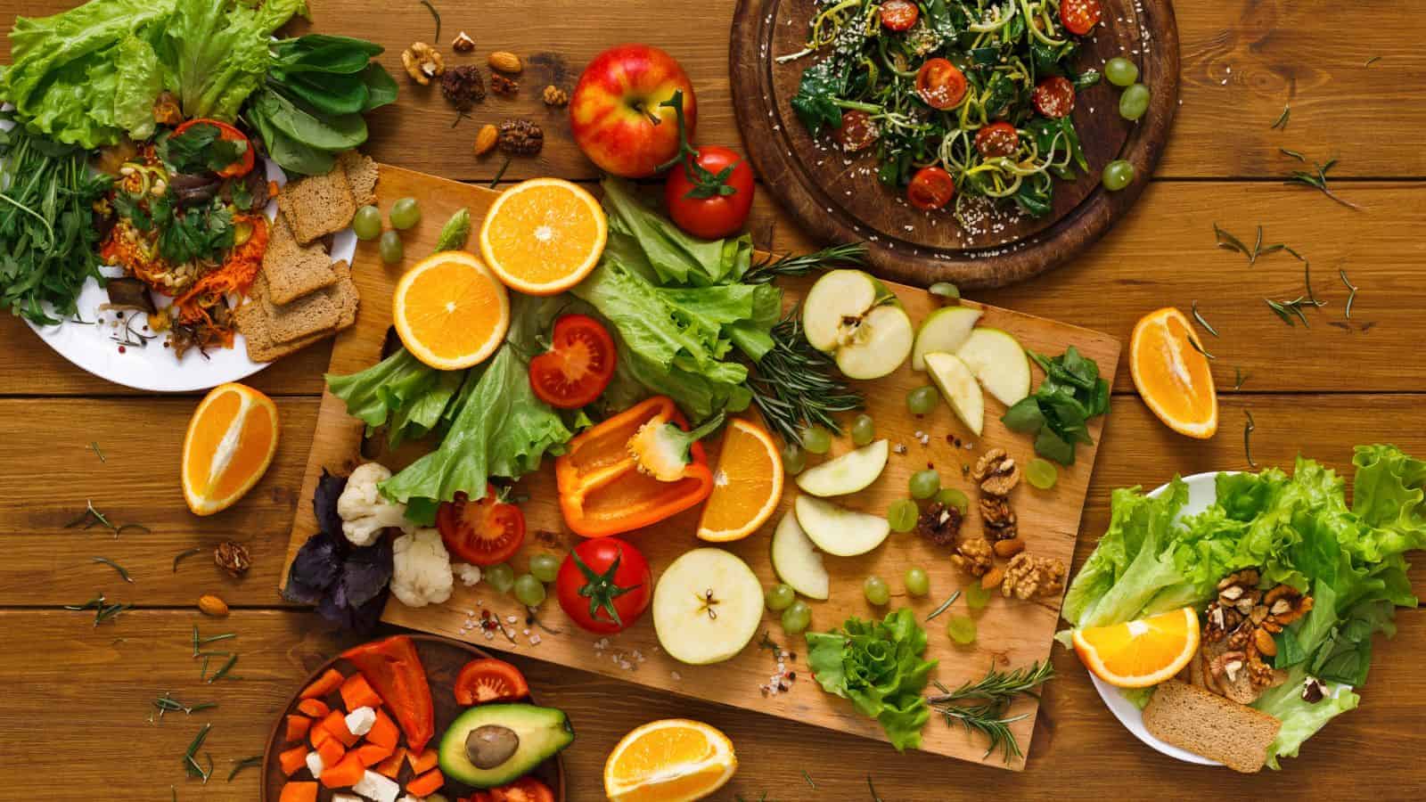 A wooden table displays an assortment of fresh fruits and vegetables, including oranges, apples, lettuce, tomatoes, avocado, and bell peppers. Nearby, drinks that increase blood sugar sit alongside plates of salad and sliced produce, garnished with nuts and herbs.