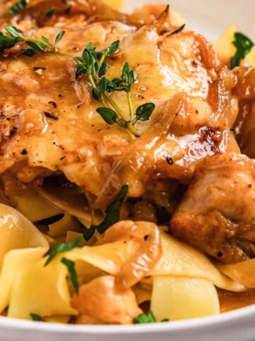 A close-up of a dish featuring wide, flat noodles topped with pieces of roasted chicken and garnished with fresh thyme. The chicken is covered in a rich, golden-brown sauce, and the noodles appear glossy and well-cooked.