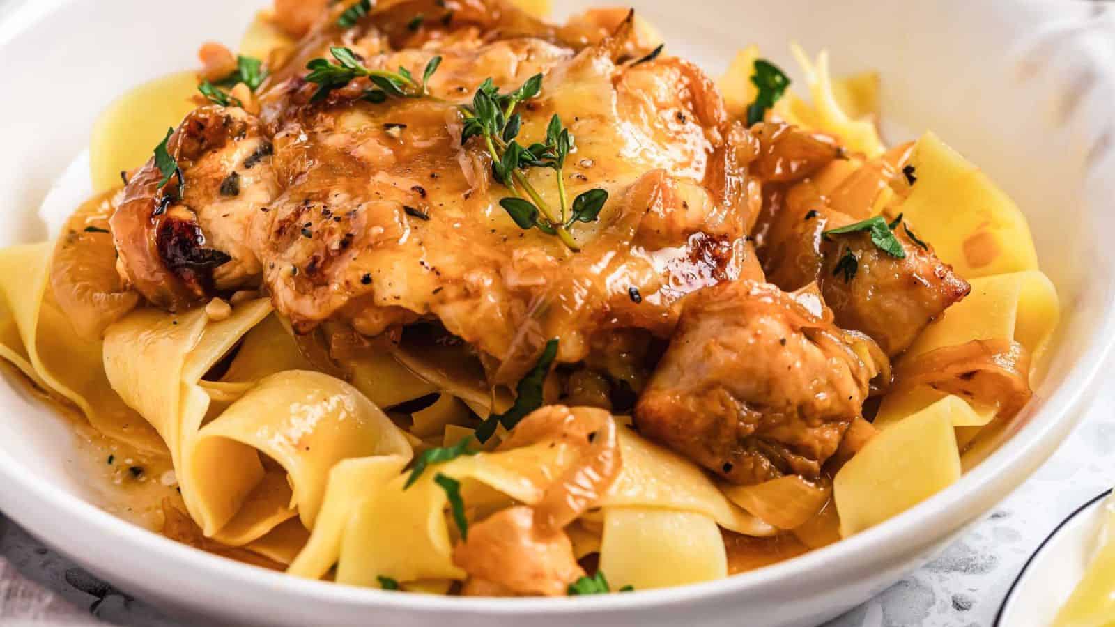 A close-up of a dish featuring wide, flat noodles topped with pieces of roasted chicken and garnished with fresh thyme. The chicken is covered in a rich, golden-brown sauce, and the noodles appear glossy and well-cooked.
