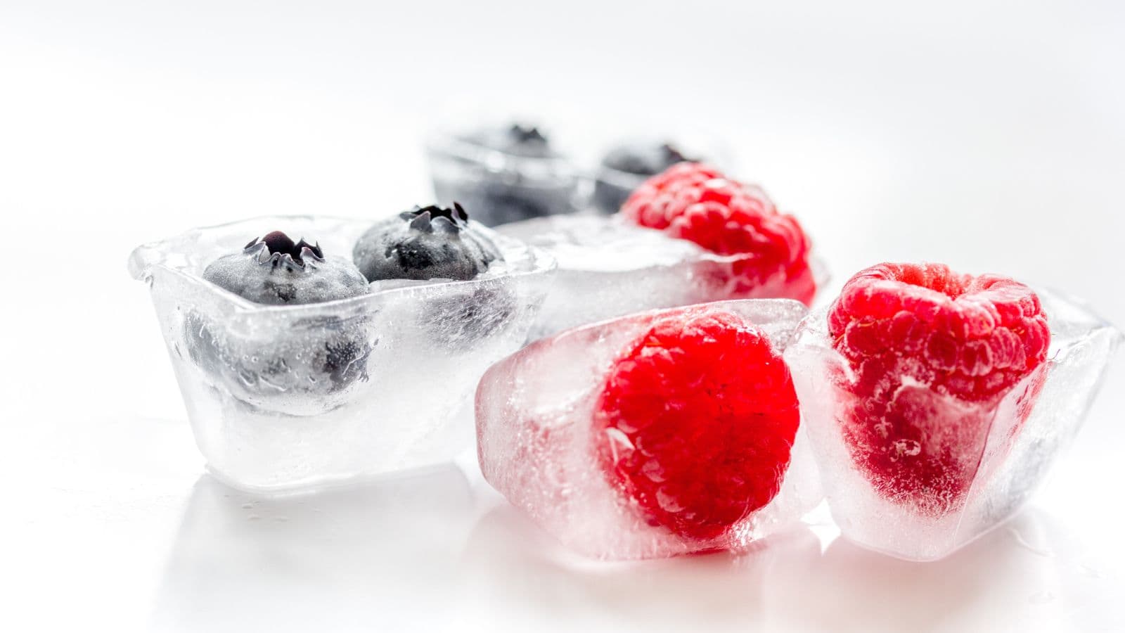 Clear ice cubes containing vibrant blueberries and raspberries, set against a white background, offering a refreshing look.
