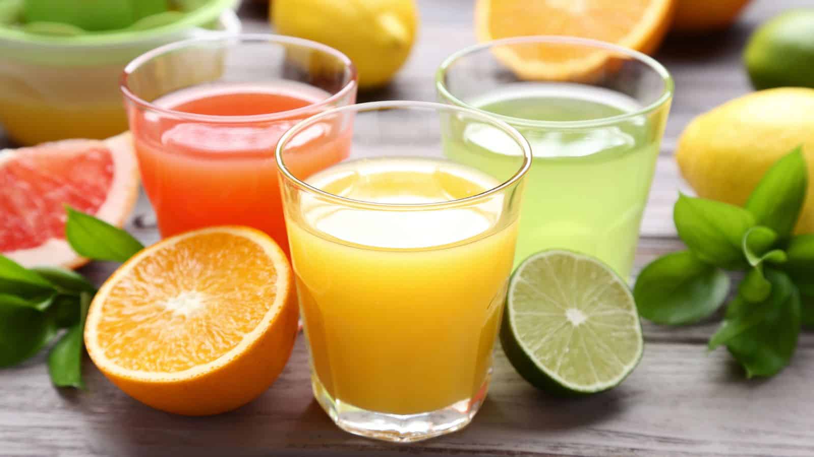 Three glasses filled with different fruit juices are arranged on a wooden surface. Slices of orange, grapefruit, and lime are placed beside them, along with scattered whole citrus fruits in the background.