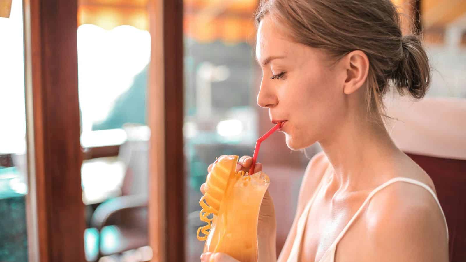 A woman sits indoors, sipping a tropical-looking drink through a red straw. The drink is garnished with a slice of mango and a spiral of peel. She is wearing a white top, and the setting includes large windows in the background.