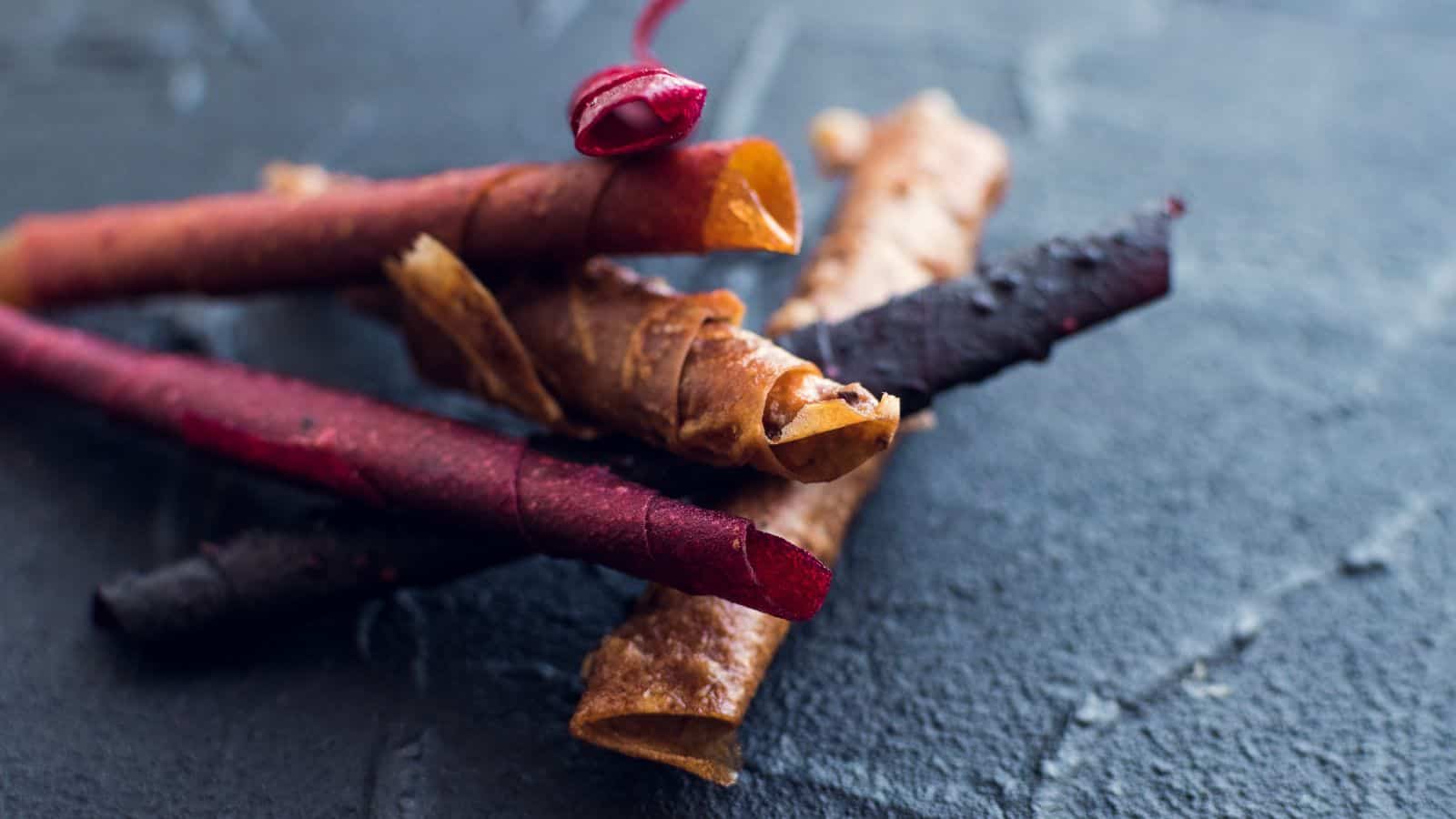 Rolled fruit leather twists in shades of red and brown, laid out on a dark textured surface, showing the rustic and chewy texture.